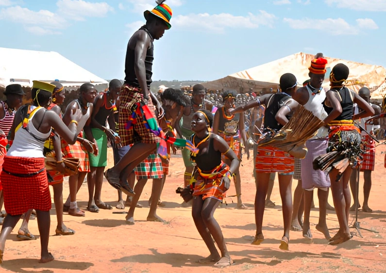 Karamojong warriors in traditional dress leaping high during a vibrant cultural dance, one man suspended mid-air against a dramatic savannah sky.