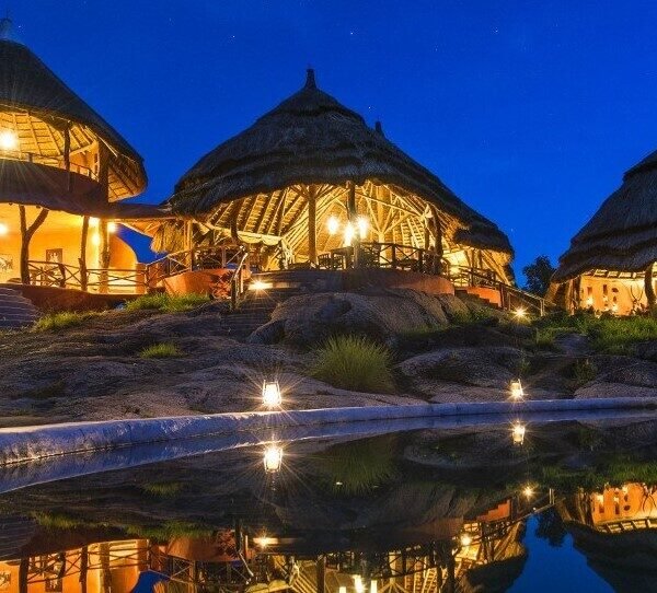 Night view of Mihingo Lodge on Lake Mburo, Uganda. Thatched-roof luxury bandas glow with warm light, reflected in the still water of a nearby pool under a starry sky.