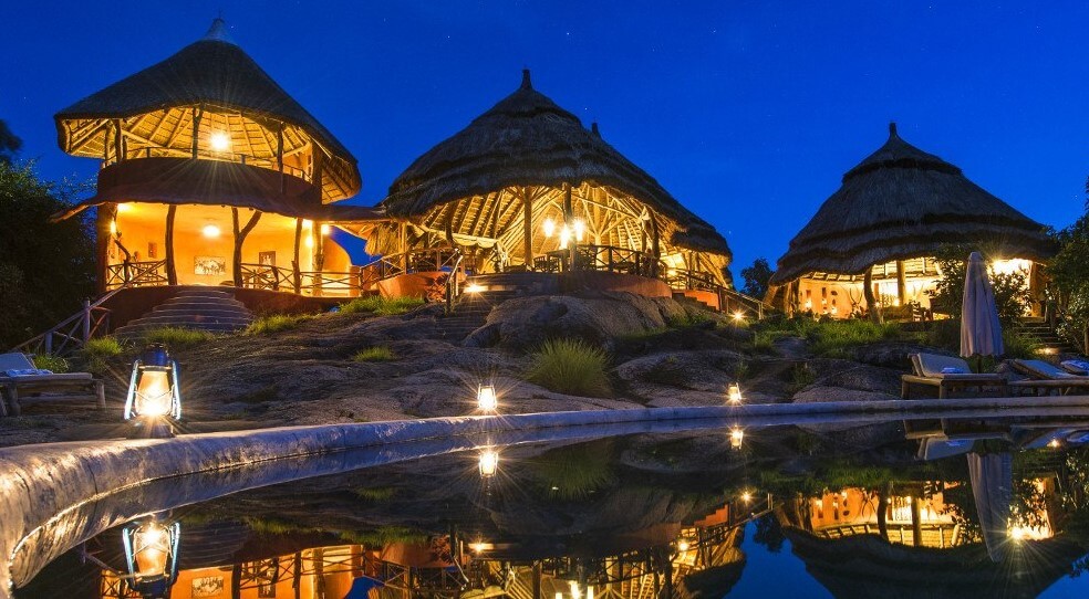 Night view of Mihingo Lodge on Lake Mburo, Uganda. Thatched-roof luxury bandas glow with warm light, reflected in the still water of a nearby pool under a starry sky.