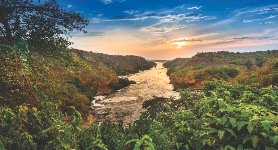 Panoramic view of the River Nile from the top of Murchison Falls at golden hour, with mist rising and sunlight casting a warm glow.