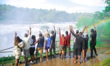 Excited tourists cheering at the top of Murchison Falls with a vivid rainbow arching in the misty background.
