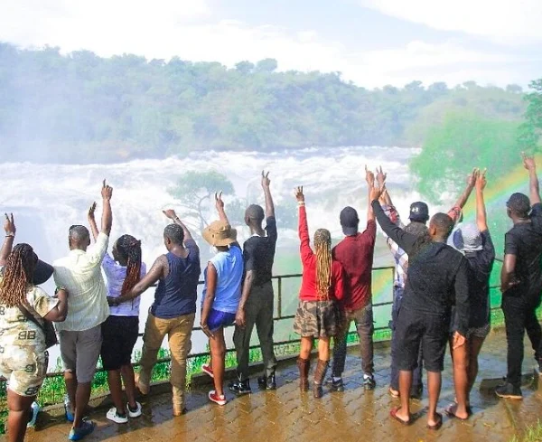 Excited tourists cheering at the top of Murchison Falls with a vivid rainbow arching in the misty background.
