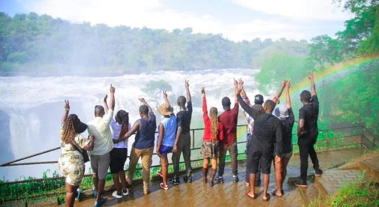Excited tourists cheering at the top of Murchison Falls with a vivid rainbow arching in the misty background.