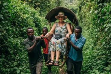 An old, fair-skinned woman, wearing a floral sundress and a wide-brimmed hat, is smiling as she is carried in a traditional wooden sedan chair. The chair is being borne on the shoulders of four strong African porters who are walking through a narrow path in a dense, vibrant green forest, typical of the Bwindi Impenetrable National Park in Uganda. The light is natural, suggesting dappled sunlight filtering through the canopy, capturing a moment from a gorilla trekking expedition.