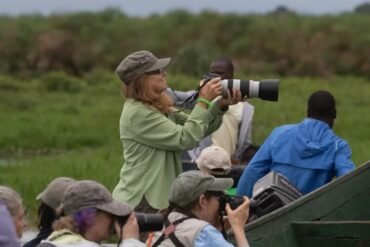 Birdwatchers in a boat on a Ugandan wetland, one person holding a large telephoto lens for bird photography