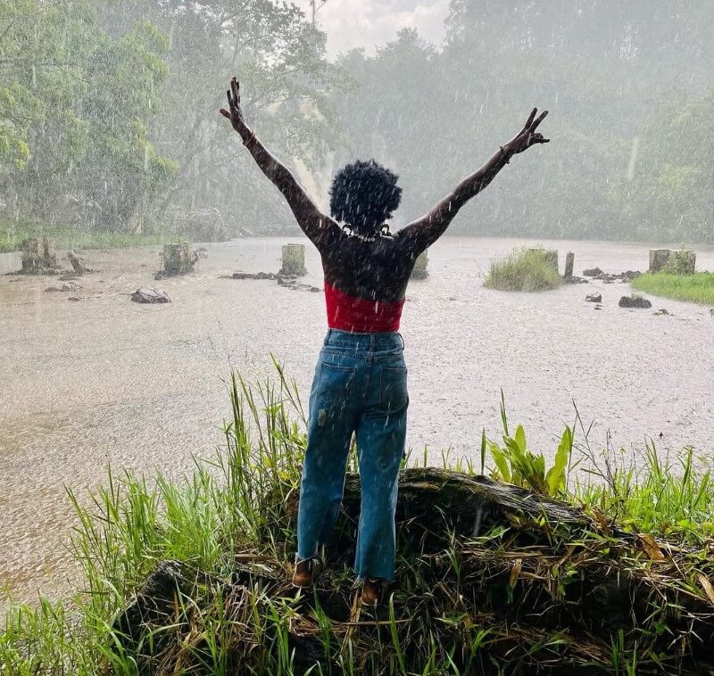 A woman standing with arms outstretched, enjoying the rain near the overflowing Ssezibwa Falls in Uganda.