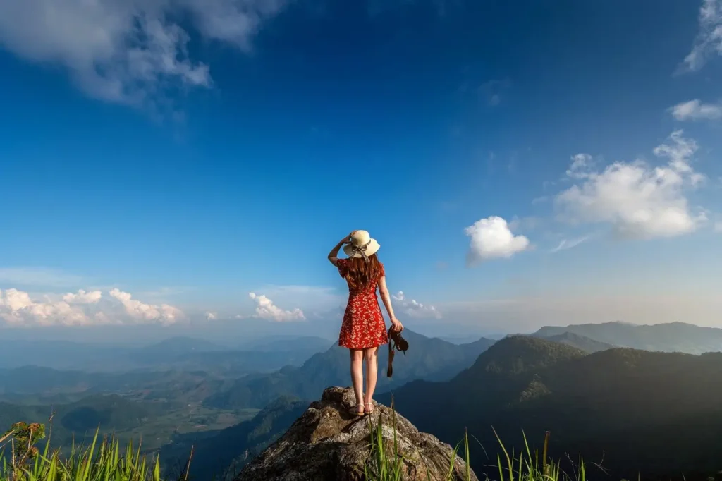 A woman in a red dress standing on a rocky mountain peak overlooking a misty tropical mountain range at sunrise in East Africa.