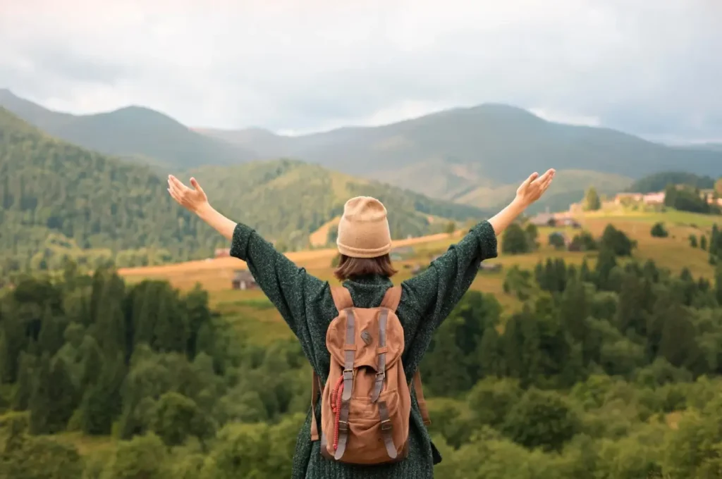 A solo traveler with a backpack standing with arms outstretched overlooking a lush, green mountain valley in Uganda.